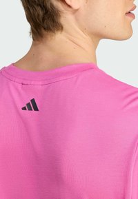 Lyserød bomulds T-shirt med rund hals og syet kant, med et sort Adidas-logo akcent på bagsiden nær halsen.