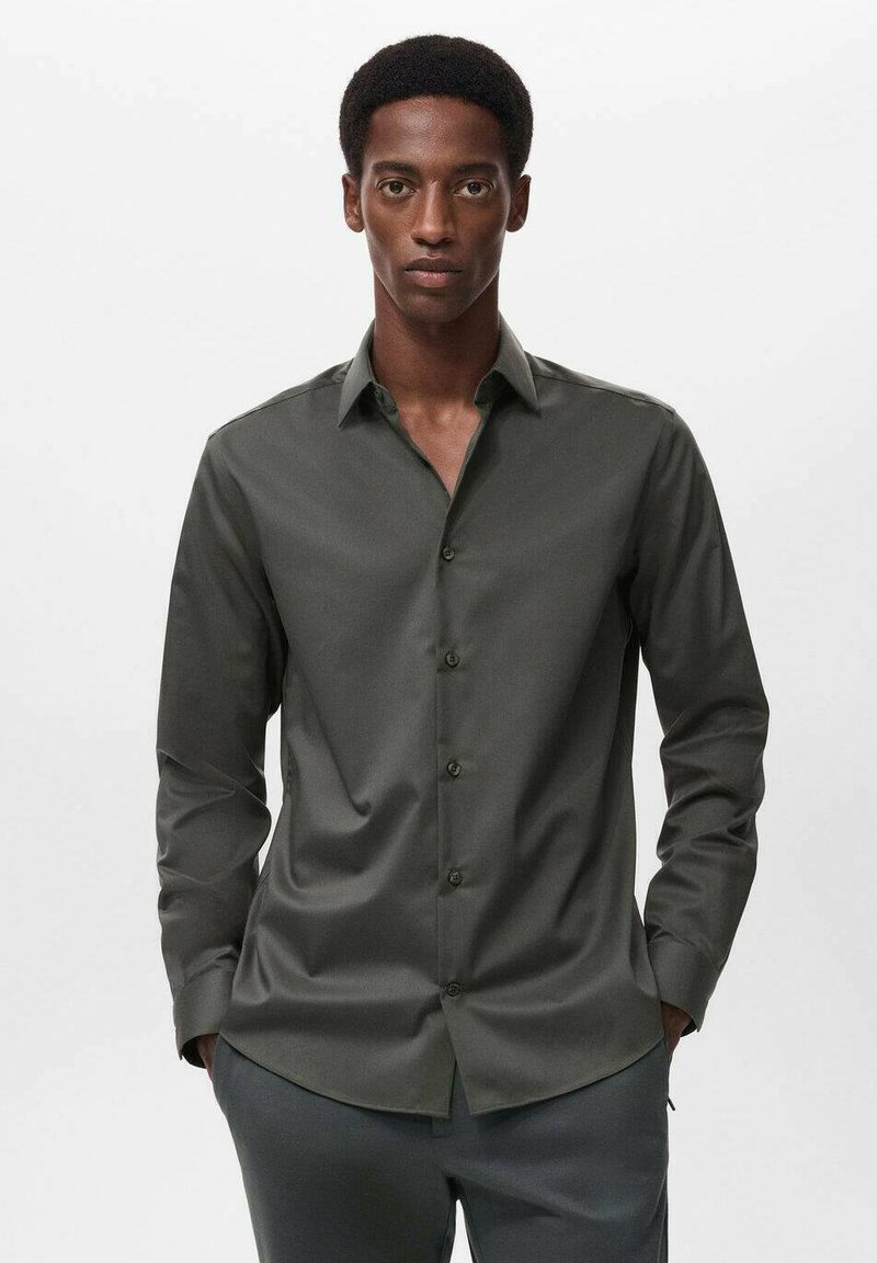 Camicia verde scuro con colletto classico, maniche lunghe, texture liscia e chiusura frontale con bottoni, indossata con pantaloni grigi.