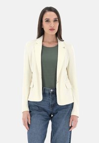 Blazer bianco con revers e due tasche frontali, abbinato a un top verde e jeans a vita alta blu. Tessuto liscio, vestibilità su misura.