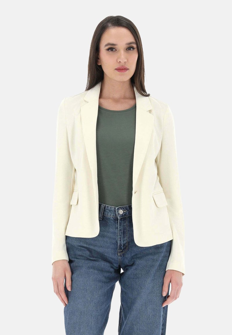 Blazer bianco con revers e due tasche frontali, abbinato a un top verde e jeans a vita alta blu. Tessuto liscio, vestibilità su misura.