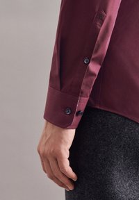 Bordeaux shirt met een knoopmanchet, voorzien van donkerblauwe knopen en een subtiele geborduurde rozenmotief nabij de schouder.