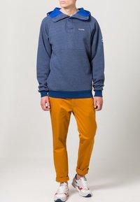 Marineblauwe fleece pullover met blauwe capuchon, kraag met twee knopen en geborduurd logo; gecombineerd met opgerolde mosterdkleurige broek en multicolor sneakers.