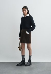 Zwarte gebreide trui, bruine suède rok met twee voorzakken, grijs gestreepte sokken en zwarte enkellaarzen, gecombineerd met een kleine handtas.