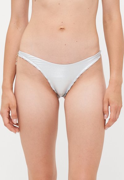 Frau trägt einen glänzenden silbernen Bikinislip mit seitlichem goldenen Charm, gezeigt von der Taille bis zur Mitte des Oberschenkels vor einem schlichten Hintergrund.