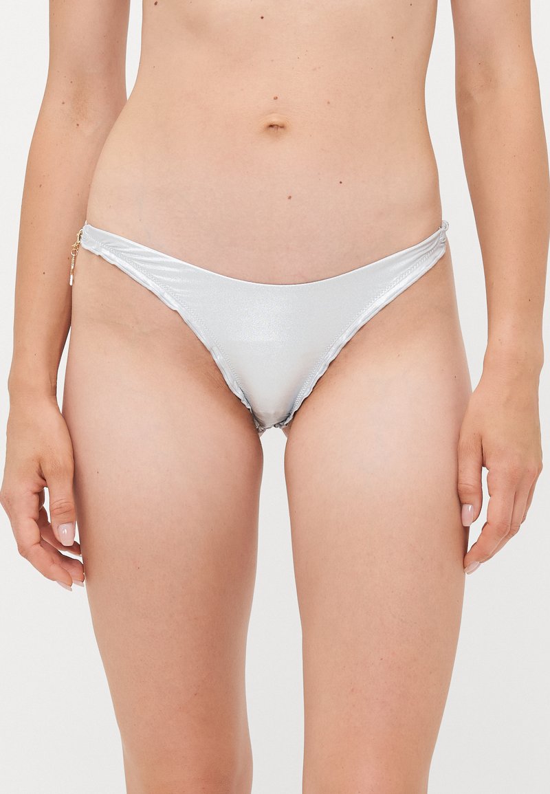 Sieviete, kas valkā spīdīgu sudraba bikini apakšdaļu ar zelta piekari, redzama no jostas līdz augšstilbam uz vienkrāsainas fona.