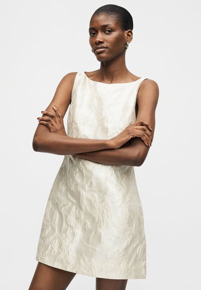 Γυναίκα με κοντά μαλλιά που φοράει ένα λευκό φόρεμα χωρίς μανίκια με υφή, όρθια με σταυρωμένα τα χέρια μπροστά σε απλό λευκό φόντο.