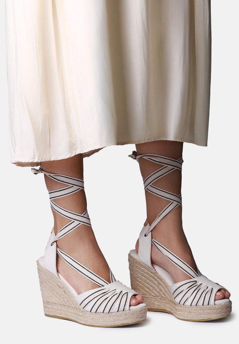 Füße tragen weiße Keilsandalen mit schwarz gestreiften, sich kreuzenden Knöchelriemchen und ein cremefarbenes Rocksaum ist darüber sichtbar.