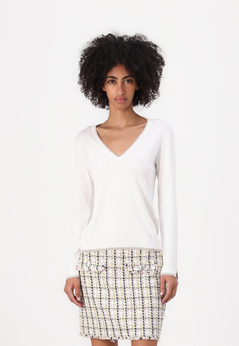 Pull-over blanc à manches longues et col en V avec une finition texturée, associé à une jupe en tweed multicolore avec des accents de boutons.