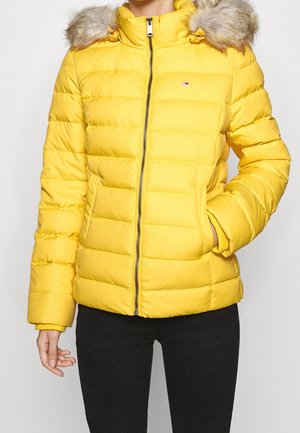 Woman wearing a bright yellow puffer jacket with fur-trimmed hood, zipped front, and black pants against a plain background.