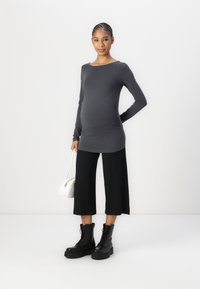 Top gris de manga larga, pantalones recortados negros de canalé y botas negras con cordones. La modelo sostiene un pequeño bolso blanco, posando frente a un fondo sencillo.