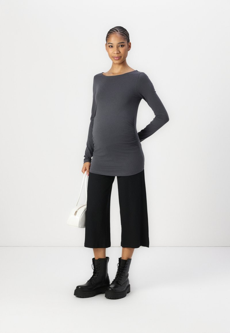 Top gris de manga larga, pantalones recortados negros de canalé y botas negras con cordones. La modelo sostiene un pequeño bolso blanco, posando frente a un fondo sencillo.