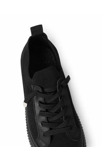 Sort sneakers med en tekstureret strikoverdel, rund tå, flade snørebånd og gummisål med en kontrastfarvet detalje nær snørehullerne.