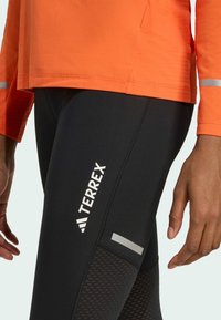 T-shirt i klar orange tyg, kombinerad med svarta leggings som har en vit "TERREX"-logotyp och reflekterande detaljer, med nätstruktur vid underbenet.