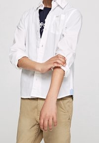 Osoba podwijająca rękaw białej koszuli z guzikami nad ciemnoniebieską koszulką, nosząca beżowe spodnie na jednolitym tle.