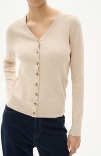Cardigan beige a coste con scollo a V, caratterizzato da sette bottoni sul davanti e maniche lunghe, abbinato a jeans blu scuro.