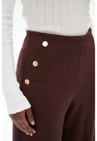Brązowe spodenki z wysokim stanem z złotymi metalowymi guzikami po bokach. Gładki materiał o strukturalnym fasonie, zestawione z białym, prążkowanym topem z długim rękawem.