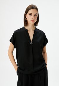 Top nero a maniche corte con scollo a V e un bottone decorativo in argento all'collo. Tessuto morbido e liscio con una vestibilità rilassata.