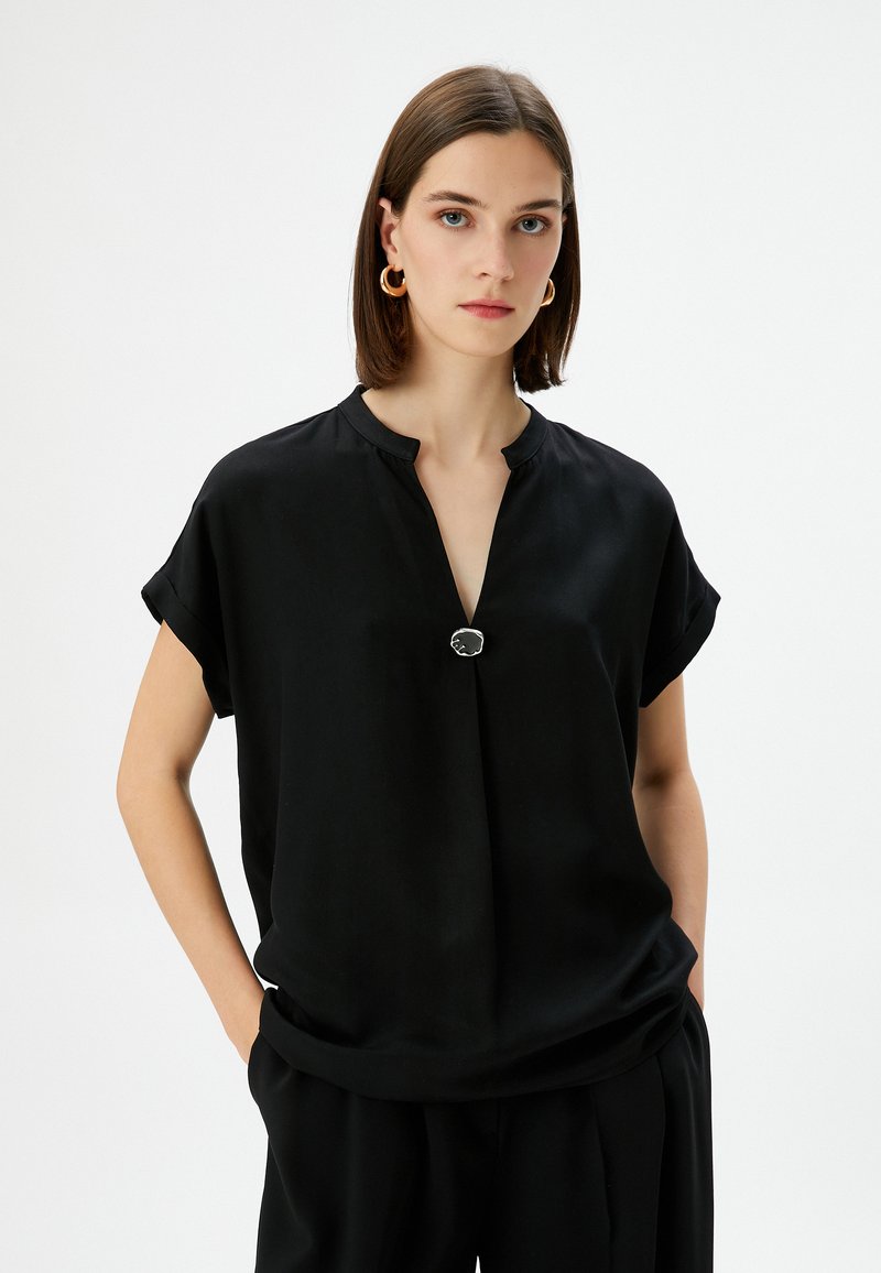 Top nero a maniche corte con scollo a V e un bottone decorativo in argento all'collo. Tessuto morbido e liscio con una vestibilità rilassata.