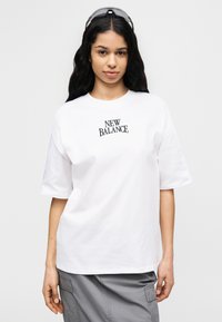 Λευκό βαμβακερό μπλουζάκι με στρογγυλή λαιμόκοψη και κοντά μανίκια. Φέρει μαύρο κείμενο "NEW BALANCE" στο στήθος. Απλός σχεδιασμός χωρίς σχέδια.