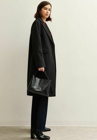 Svart ullkappa med skräddarsydd design, kombinerad med svarta högklackade stövlar. Håller en strukturerad svart läderhandväska. Mörkblå vida jeans.