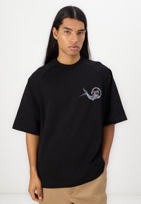 Černé oversized tričko vyrobené z bavlny s raglánovými rukávy, se barevným vyšitým designem ryby a textem na levé straně hrudi.