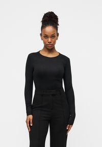 Μαύρο μακρυμάνικο μπλουζάκι από ελαστικό ύφασμα, με στρογγυλή λαιμόκοψη και εφαρμοστή γραμμή, συνδυασμένο με μαύρο παντελόνι ψηλόμεσο.