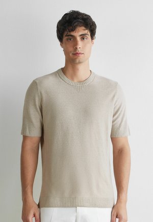 Ung mand med kort krøllet hår iført en beige kortærmet striktrøje og hvide bukser, stående mod en ensartet lys baggrund.