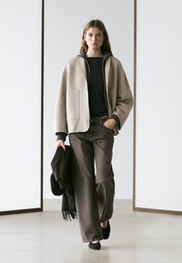 Frau geht drinnen und trägt eine beige übergroße Jacke, einen dunklen Pullover, weite Jeans, hält in einer Hand einen dunklen Schal und eine Tasche.