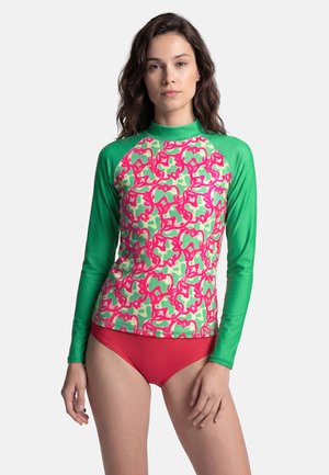 Woman wearing a long-sleeve green rash guard with pink and light green abstract patterns and solid red swim bottoms, standing against a white background.