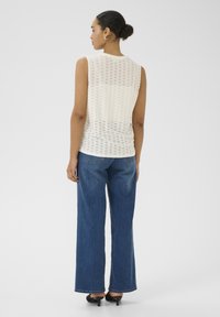Haut sans manches en tricot crème avec un motif texturé. Associé à un jean bleu taille haute. Le mannequin est de dos, montrant le design au dos.