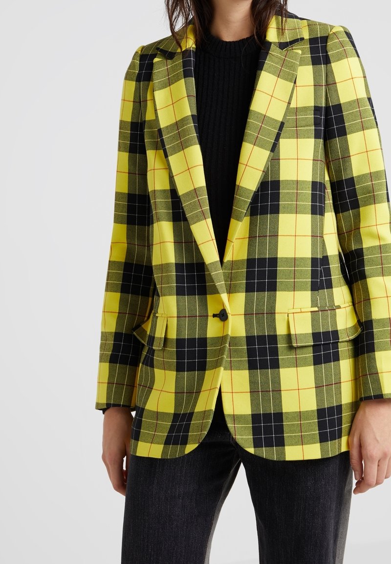 Blazer à carreaux jaune et noir avec une coupe ajustée, un bouton unique et deux poches avant, porté sur un pull noir texturé.