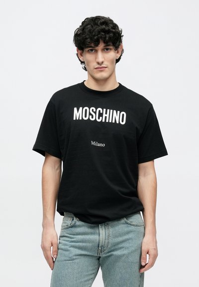 Juoda medvilninė marškinėliai su ryškiu baltu užrašu „MOSCHINO“ ir po juo esanti „Milano“. Modelis dėvi šviesiai mėlynas džinsines kelnes, stovėdamas prieš paprastą foną.