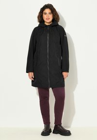 Abrigo negro impermeable con capucha, cierre con cremallera y bolsillos laterales, combinado con pantalones ajustados burdeos y botas negras de tobillo. Diseño sencillo.