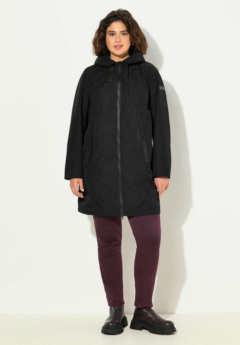 Abrigo negro impermeable con capucha, cierre con cremallera y bolsillos laterales, combinado con pantalones ajustados burdeos y botas negras de tobillo. Diseño sencillo.