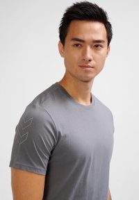 Grijze t-shirt met korte mouwen en een gladde textuur. Heeft een ronde hals en drie chevron-patronen in een lichtere tint op de linkerschouder.