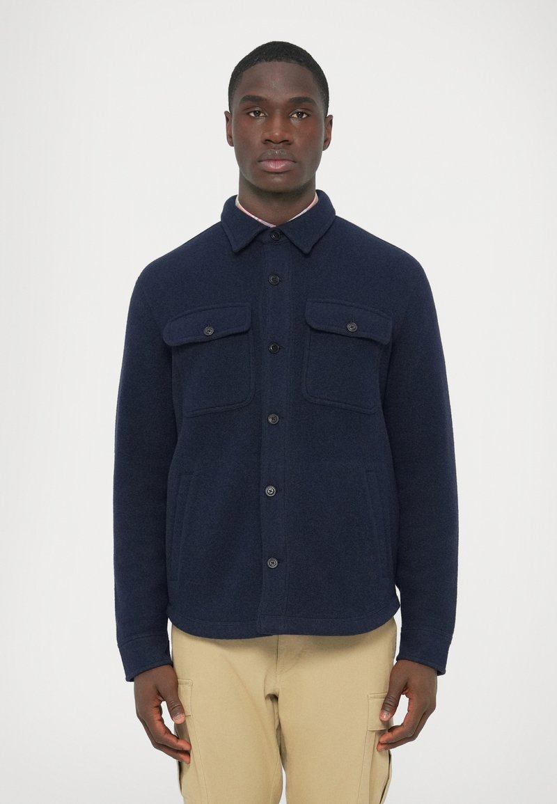 Casaco-roupa de lã azul-marinho com dois bolsos frontais no peito, fecho de botões e gola com colarinho. Tecido texturizado com um corte descontra�ído.