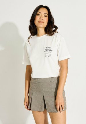 Mujer de pie vistiendo una camiseta blanca con texto negro e ilustración de perro, combinada con una falda de cuadros plisada de talle alto.
