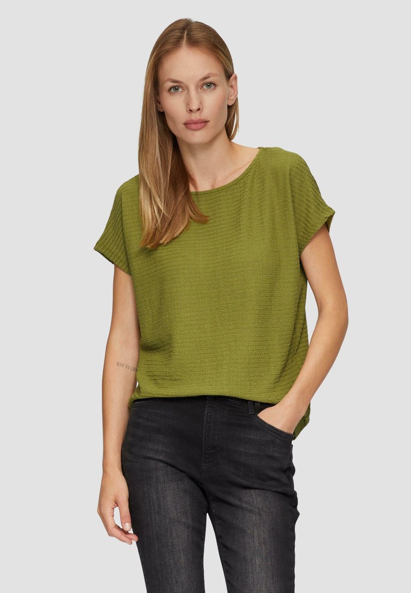 Groene textuur korte mouw top met een relaxte pasvorm, gecombineerd met donkere jeans. De stof heeft een subtiel, golvend patroon. Eenvoudig ontwerp zonder zichtbare accenten.
