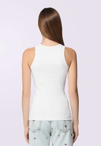 Donna con lunghi capelli castani lisci, che indossa un top bianco a coste e jeans azzurri chiari con borchie argentate, in piedi davanti a uno sfondo chiaro.