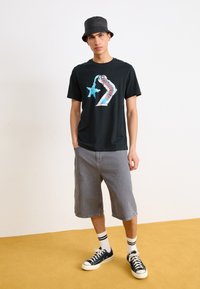 Camiseta negra con un gráfico colorido, pantalones cortos grises, gorra de cubo negra y zapatillas azul marino con suelas blancas y calcetines a rayas.