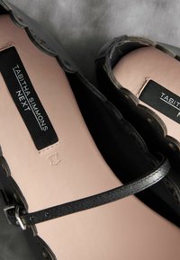 Le calzature in pelle nera presentano un bordo scallopato, una sottile cinghia alla caviglia e una suola interna beige liscia con dettagli del marchio.