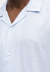 Φωτεινό γαλάζιο λινό πουκάμισο με κοντό γιακά και δύο λευκά κουμπιά, φορεμένο από άτομο με σκούρο δέρμα ορατό στον λαιμό.