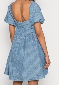 Robe en denim bleu avec un motif rayé texturé, manches courtes à volants, et un décolleté en V profond, présentant une taille froncée et une jupe évasée.