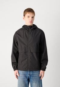 Zwarte windbreaker jas met geometrische textuur, ritssluiting aan de voorkant, verstelbare capuchon en elastische manchetten, gecombineerd met lichtblauwe jeans.