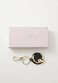 Black keychain made of textured leather with gold hardware, featuring a round shape, clasp, and chain detail, presented in a pink box.