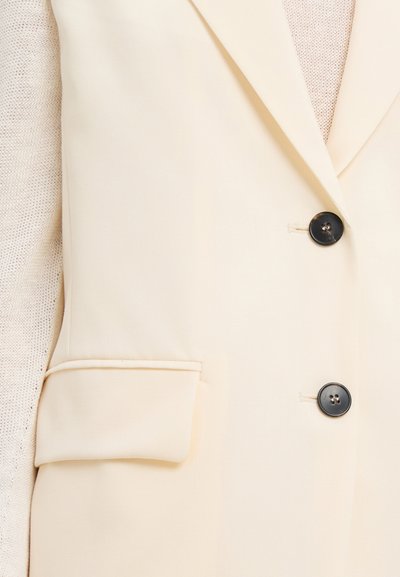 Beige skræddersyet blazer med en struktureret design, der har en enkelt brystlomme, to sorte knapper og en glat stoftekstur.