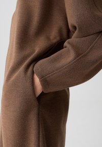 Bruin stoffen kledingstuk met een gestructureerde oppervlakte, met een losse pasvorm en zijzakken, zichtbaar bij de mouw- en zakopening.