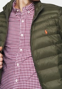 Olive green puffer jacket with zip, featuring horizontal quilted design. Red and white checkered button-down shirt underneath.