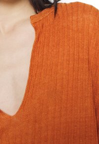 Lähellä otettu kuva henkilöstä, joka on pukeutunut ribbittyyn, oranssiin neulepaitaan, jossa on syvä V-pääntie, ja tummat hiukset näkyvät osittain.