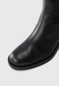 Μαύρη δερμάτινη μποτάκια αστράγαλου με λεία υφή, στρογγυλή μύτη και μινιμαλιστικό σχέδιο. Διαθέτει καουτσούκ σόλα και διακριτές ραφές.